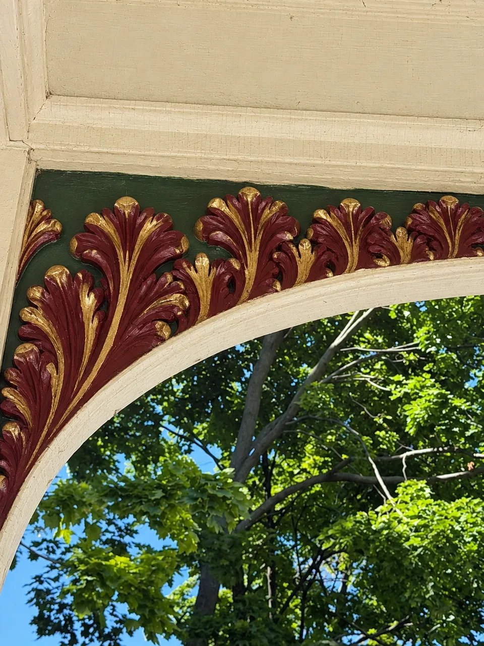The porch arch as it will look painted: burgundy acanthus leaves edged in gold on a deep green ground under cream trim, with trees and sky beyond.